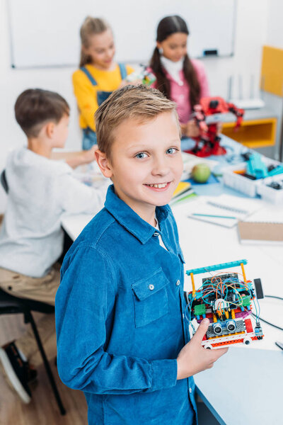smiling schoolboy holding robot and looking at camera with classmates at background in classroom