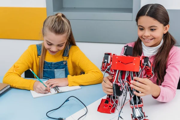 Alunas Sorridentes Escrevendo Caderno Segurando Robô Vermelho Mesa Aula Educação — Fotografia de Stock