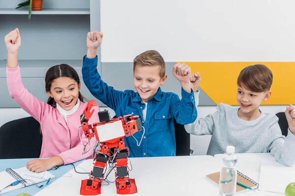 happy schoolchildren raising hands and looking at red handmade robot at desk during STEM lesson