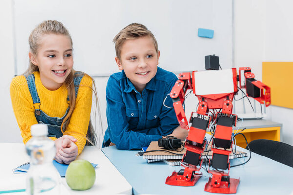 Улыбающиеся школьники сидят за столом и смотрят на красного пластикового робота в классе стеблей

