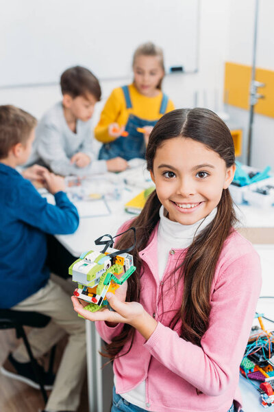 smiling schoolgirl holding robot and looking at camera with classmates at background