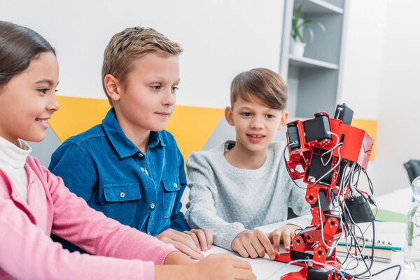 smiling schoolchildren looking at red plastic robot in classroom