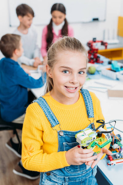 preteen schoolgirl holding multicolored robot and looking at camera in classroom