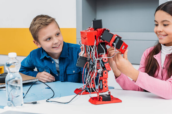 adorable schoolgirl and smiling schoolboy sitting at desk and working together on robot model at STEM class