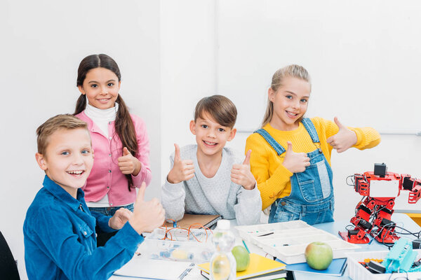 cheerful schoolchildren looking at camera and showing thumbs up while having STEM lesson