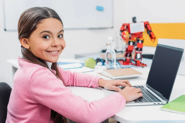 可爱的女学生坐在桌子上与机器人模型 看着相机和使用笔记本电脑与空白屏幕在干细胞课 — 图库照片