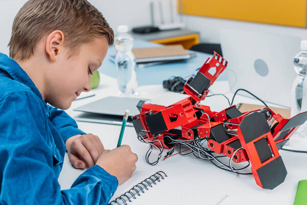 schoolboy sitting at desk with robot model and writing in notebook during STEM lesson
