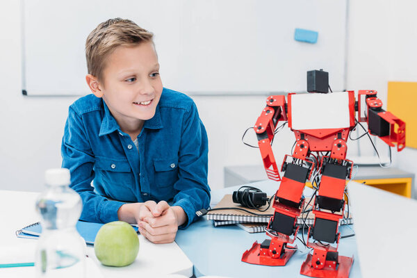 smiling schoolboy sitting at table and looking at robot model during STEM lesson  