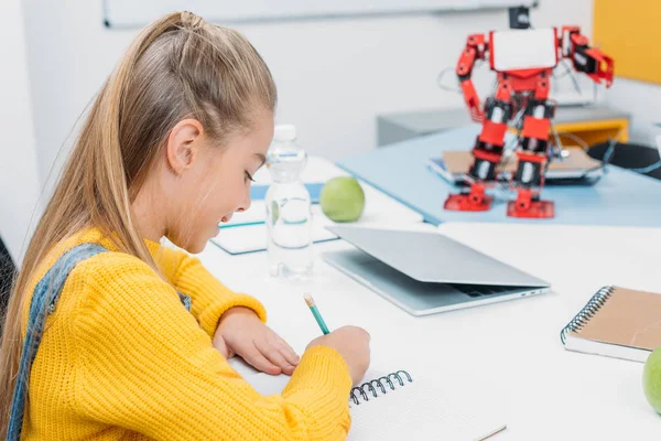 schoolgirl sitting at desk with robot model and writing in notebook during STEM lesson