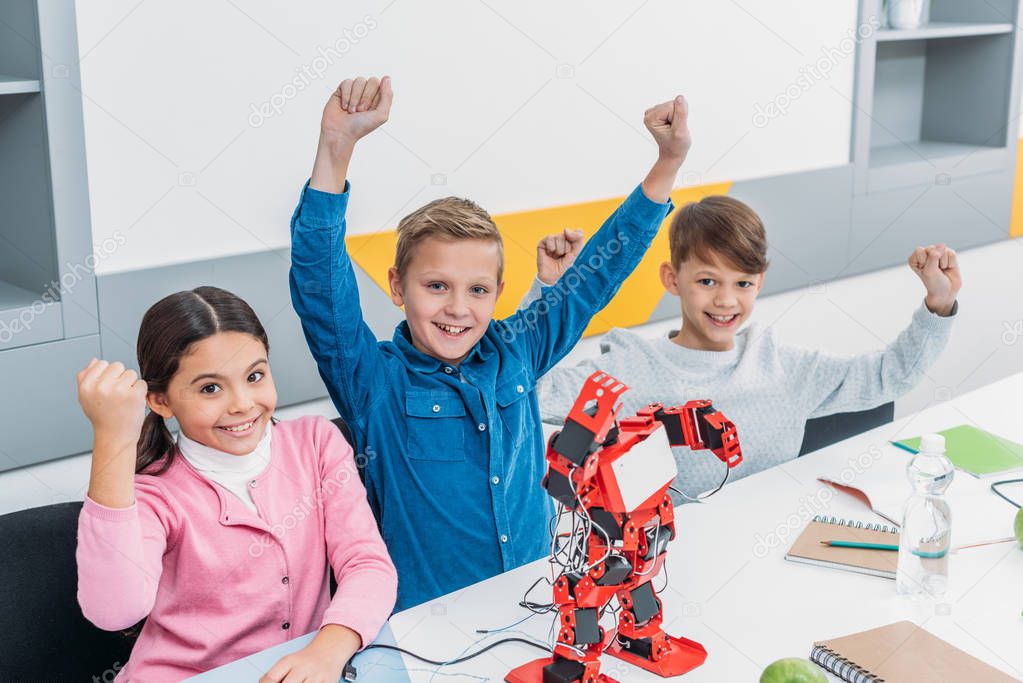 children joying and raising hands at desk with electric robot during stem lesson
