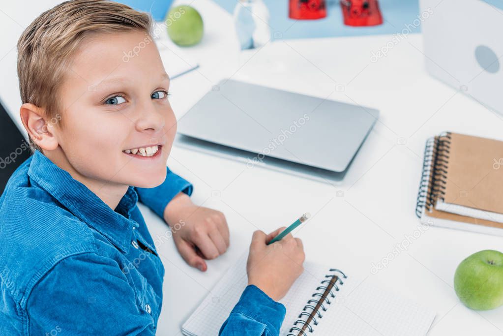 smiling boy writing in notebook, smiling and looking at camera in stem class