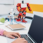 Cropped view of schoolgirl sitting at table with robot model and using laptop with blank screen during STEM lesson