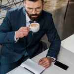 Hochwinkelaufnahme des bärtigen Geschäftsmannes in Brille, der am Arbeitsplatz Kaffee trinkt