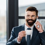 Sonriente hombre de negocios barbudo sosteniendo una taza de café y hablando por teléfono inteligente