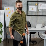 Knappe bebaarde zakenman in brillen houden skateboard en lachend op camera in kantoor