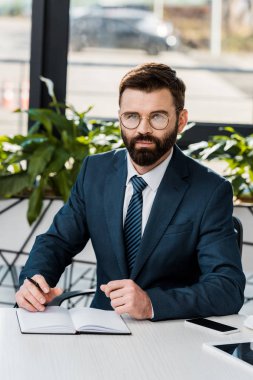 kendinden emin buisnessman gözlük sittting işyerinde ve kameraya arıyor