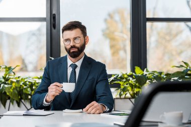 takım elbise ve kahve tutarak ve kamera Office bakarak gözlük ciddi sakallı işadamı