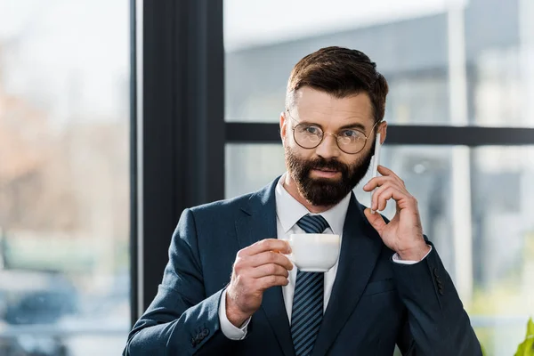 专业的胡须商人拿着一杯咖啡 在办公室用智能手机说话 — 免费的图库照片