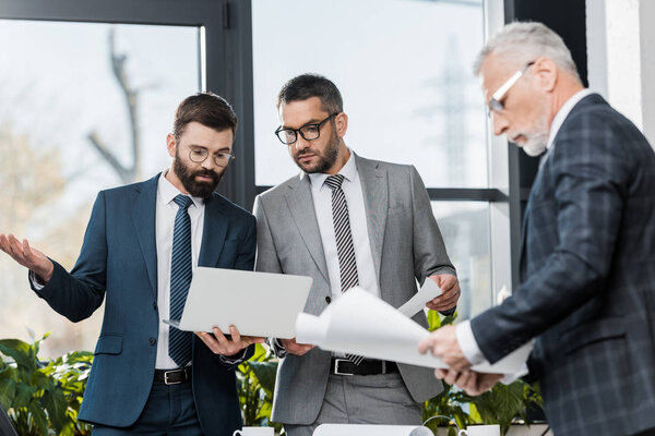 три серьезных профессиональных бизнесмена, работающих с документами и ноутбуком в офисе
