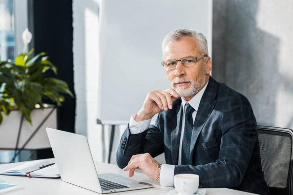 confident mature businessman using laptop and looking at camera while sitting at workplace