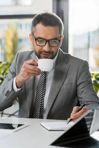 Affärsman Kostym Och Glasögon Dricker Kaffe Och Skriver Anteckningsboken Wokrplace — Gratis stockfoto