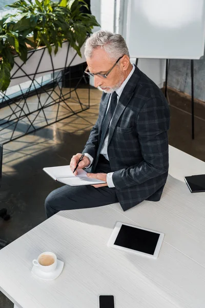 成熟したビジネスマンのオフィスのテーブルの上に座ってしながらノートに書いてのハイアングル  — 無料ストックフォト