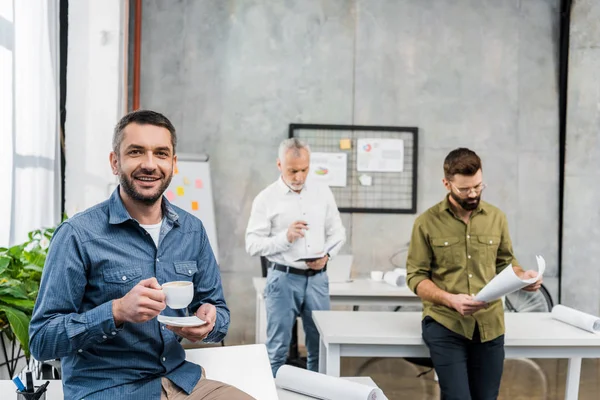 Bonito Empresário Com Xícara Café Sorrindo Para Câmera Enquanto Colegas — Fotos gratuitas