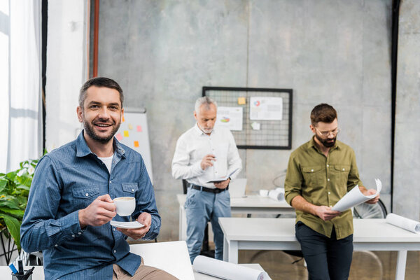 handsome businessman with cup of coffee smiling at camera while colleagues woking behind
