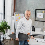 Smiling handsome middle aged businessman standing with longboard in office