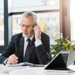 Gutaussehender Geschäftsmann mittleren Alters sitzt im Büro am Tisch und unterhält sich per Smartphone