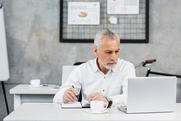 serious handsome middle aged businessman holding pen and looking at laptop in office