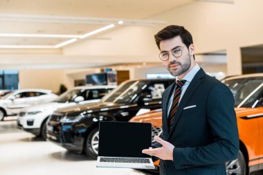 happy male car dealer in eyeglasses pointing at laptop with blank screen in car salon clipart