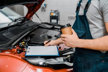partial view of male auto mechanic with notepad and coffee to go near car at auto repair shop clipart