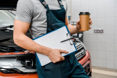 partial view of male auto mechanic with notepad and coffee to go near car at auto repair shop clipart