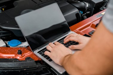 partial view of auto mechanic working on laptop with blank screen at automobile with opened car cowl at mechanic shop clipart