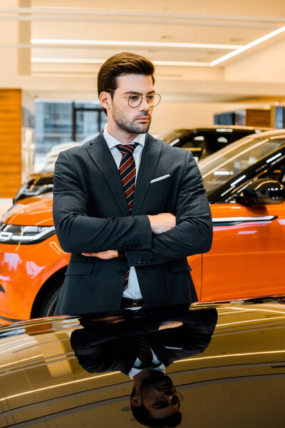 serious businessman in eyeglasses posing with crossed arms near black automobile