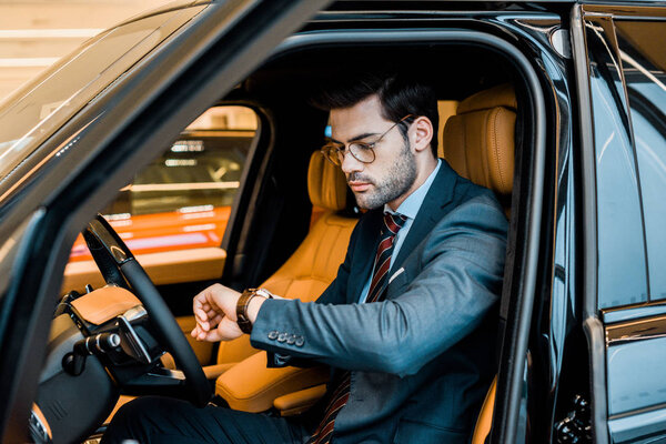 stylish businessman in eyeglasses checking watch in luxury car