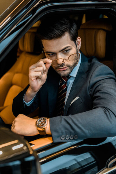 stylish businessman with luxury watch putting on eyeglasses while sitting in car