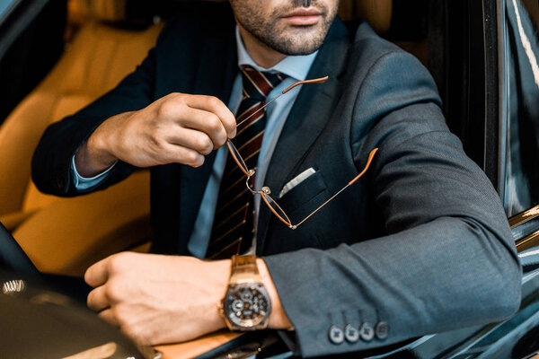 partial view of businessman with luxury watch holding eyeglasses while sitting in car