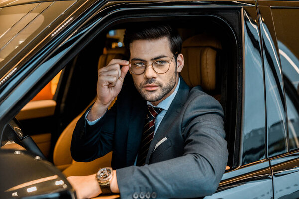 young businessman with luxury watch putting on eyeglasses while sitting in car