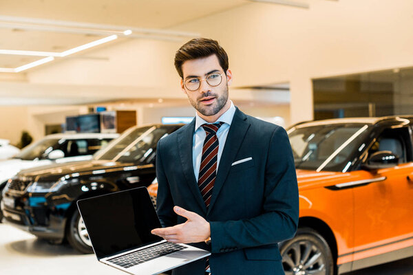 smiling male car dealer in eyeglasses pointing at laptop with blank screen in car salon
