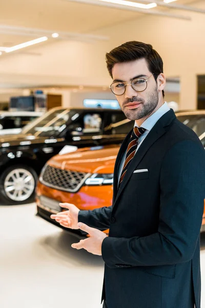 Junger Männlicher Autohändler Mit Brille Zeigt Autosalon Mit Den Händen — Stockfoto