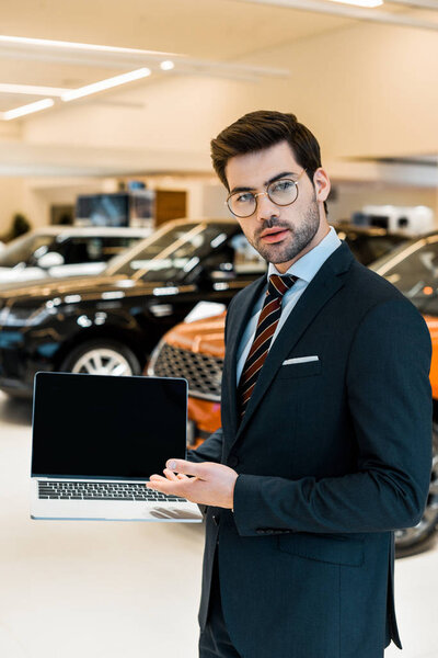 young male car dealer in eyeglasses pointing at laptop with blank screen in car salon