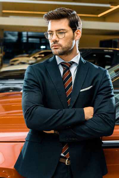 serious businessman in eyeglasses posing with crossed arms near automobile 