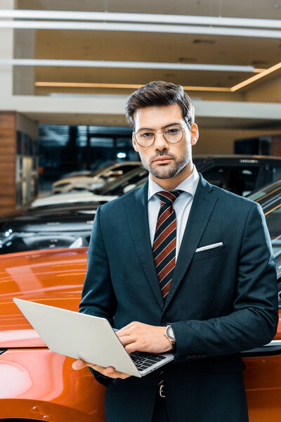 stylish young businessman in eyeglasses using laptop near automobile 