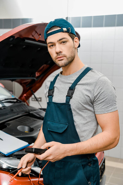 портрет автомеханика, держащего мультиметр вольтметр для проверки напряжения аккумулятора в автомастерской
