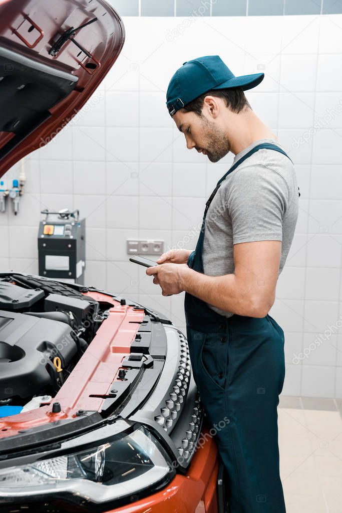 auto mechanic in uniform using smartphone at car with opened cowl at mechanic shop