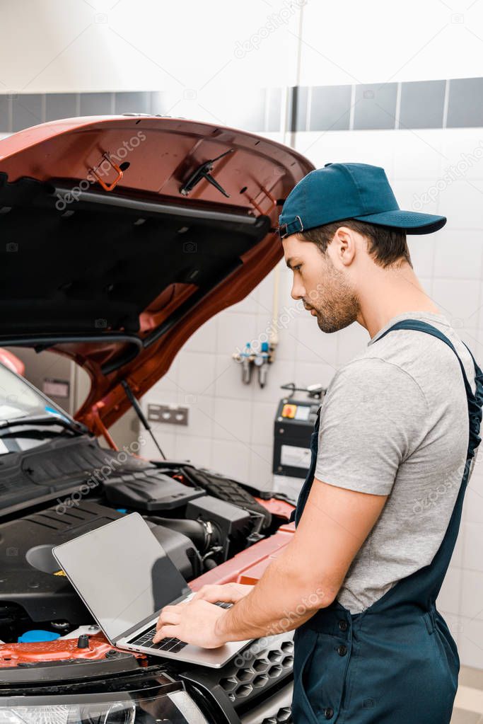 side view of auto mechanic working on laptop at automobile with opened car cowl at mechanic shop