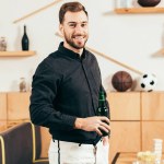 Retrato del hombre sonriente con botella de cerveza de pie en la cafetería con futbolín