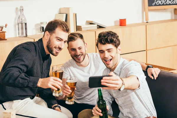 portrait of smiling friends with beer taking selfie on smartphone together in cafe